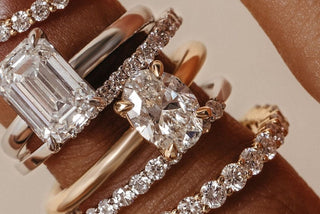Close-up of stacked diamond rings on a finger, featuring an emerald cut and an oval cut center stone with pavé and plain gold bands, classic, elegant, and full of sparkle.
