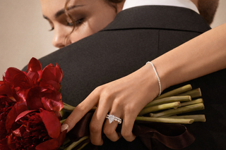 Close-up of a woman holding deep red peonies while wearing a pear-shaped diamond engagement ring with matching band and delicate tennis bracelet