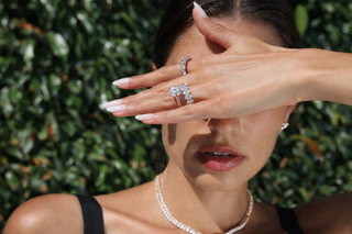 Woman covering her eyes in sunlight wearing stacked diamond rings and a delicate tennis necklace against a green foliage background