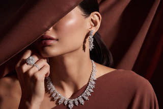 Woman wearing layered diamond necklace with matching drop earrings and a diamond ring styled against a rich brown fabric backdrop
