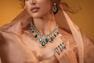 Model wearing an emerald and diamond high jewelry necklace with matching earrings and ring against a warm gold backdrop