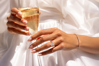 Elegant close-up of a woman holding a champagne flute while showcasing a radiant-cut diamond engagement ring in a luxurious bridal setting.