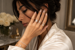 Elegant woman wearing multiple diamond and gold rings with a diamond tennis bracelet, posed in a luxurious interior with soft lighting and neutral tones.