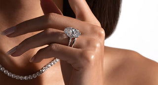 Close-up of a woman wearing an oval diamond engagement ring in white gold with a delicate diamond tennis necklace in the background