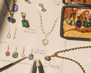 Flat lay of custom diamond and gemstone jewelry sketches with hand-drawn design notes, colored stone pendants, gold settings, and jewelry tools on a designer’s workspace