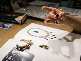 A hand holds a teardrop earring over an open book with jewelry designs. Items include a necklace and bracelet. The setting feels elegant and artistic.
