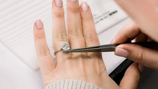 A jeweler uses tweezers to adjust a round brilliant-cut diamond engagement ring on a woman's finger, showcasing the stone's clarity and sparkle against a soft, light background.