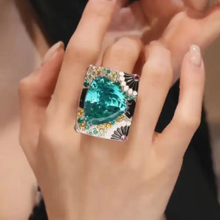 A luxurious silver statement ring in a middle finger featuring a large trillion-cut teal Paraiba gemstone at the center. The wide band is intricately adorned with pink, orange, green, and white gemstones in floral and geometric patterns, showcasing elaborate craftsmanship and vibrant color.