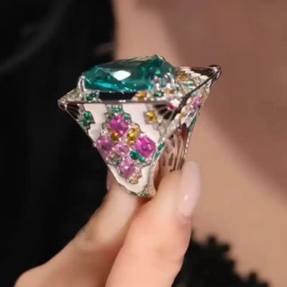 A luxurious silver statement ring  featuring a large trillion-cut teal Paraiba gemstone at the center. The wide band is intricately adorned with pink, orange, green, and white gemstones in floral and geometric patterns, showcasing elaborate craftsmanship and vibrant color. in between holding in the index fingure and thumb.
